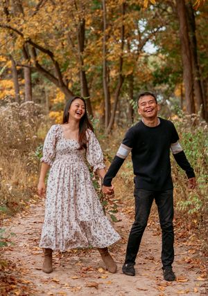 A couple walks hand in hand through a wooded path filled with golden autumn foliage on a sunny afternoon.