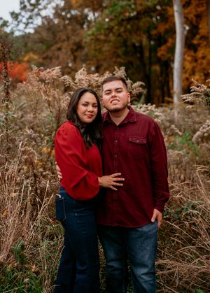A couple in matching burgundy sweaters pose together among tall autumn grasses during a fall photo session.