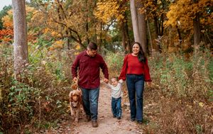 A family walks hand in hand down a scenic autumn trail surrounded by golden foliage and fallen leaves.