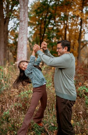 A couple dances playfully together in a fall forest setting with brown foliage and tall trees in the background.