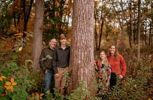 People pose together in a wooded autumn setting with tall trees and fallen leaves in the background.
