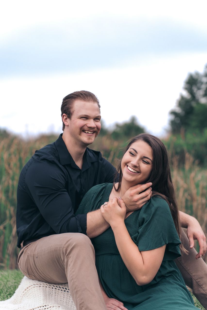 A happy couple sits close together on a blanket outdoors, sharing a candid laughing moment in a natural setting. Marion, Ohio