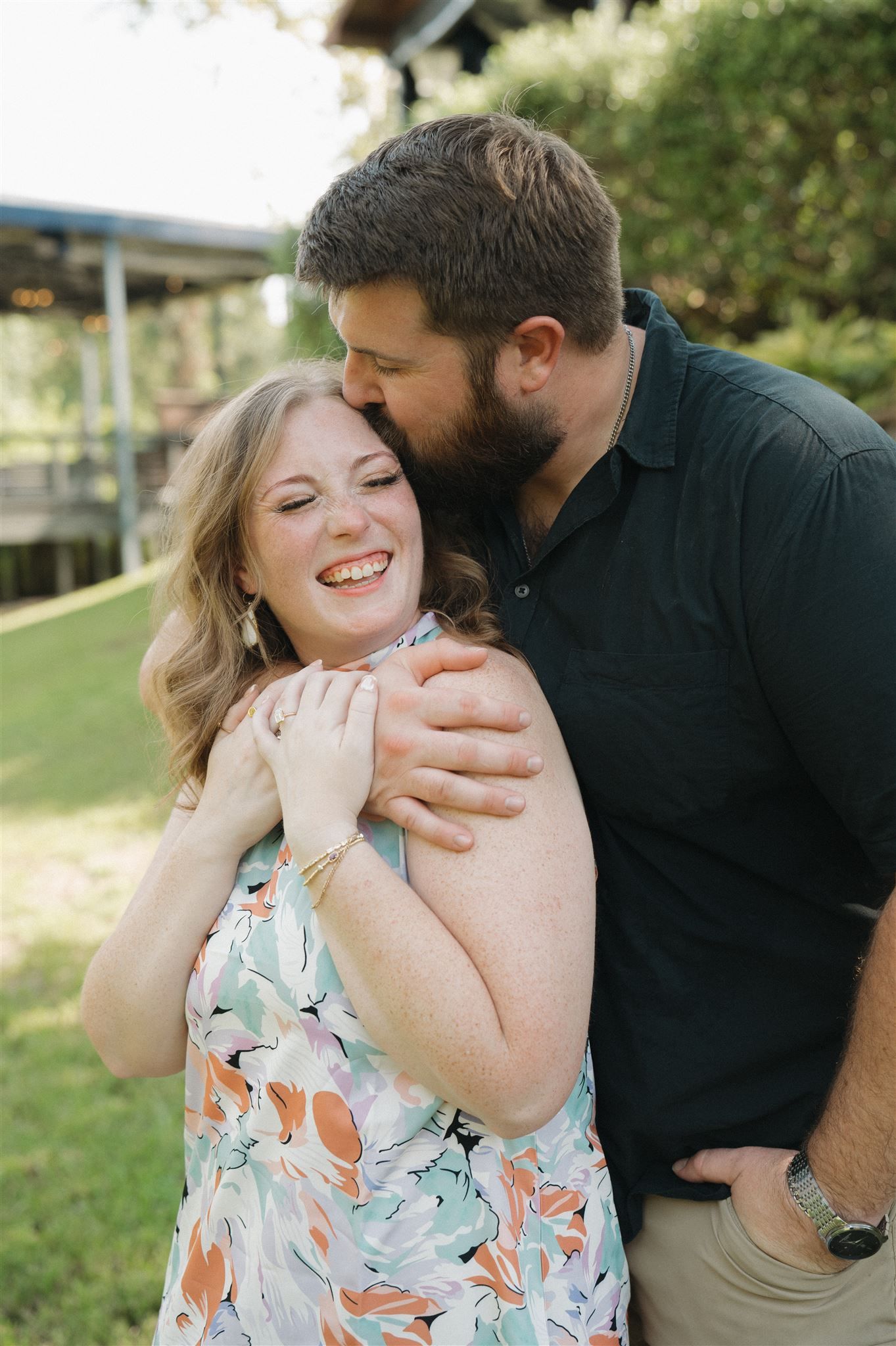 A joyful couple embraces and laughs together outdoors on a sunny day, with greenery in the background.