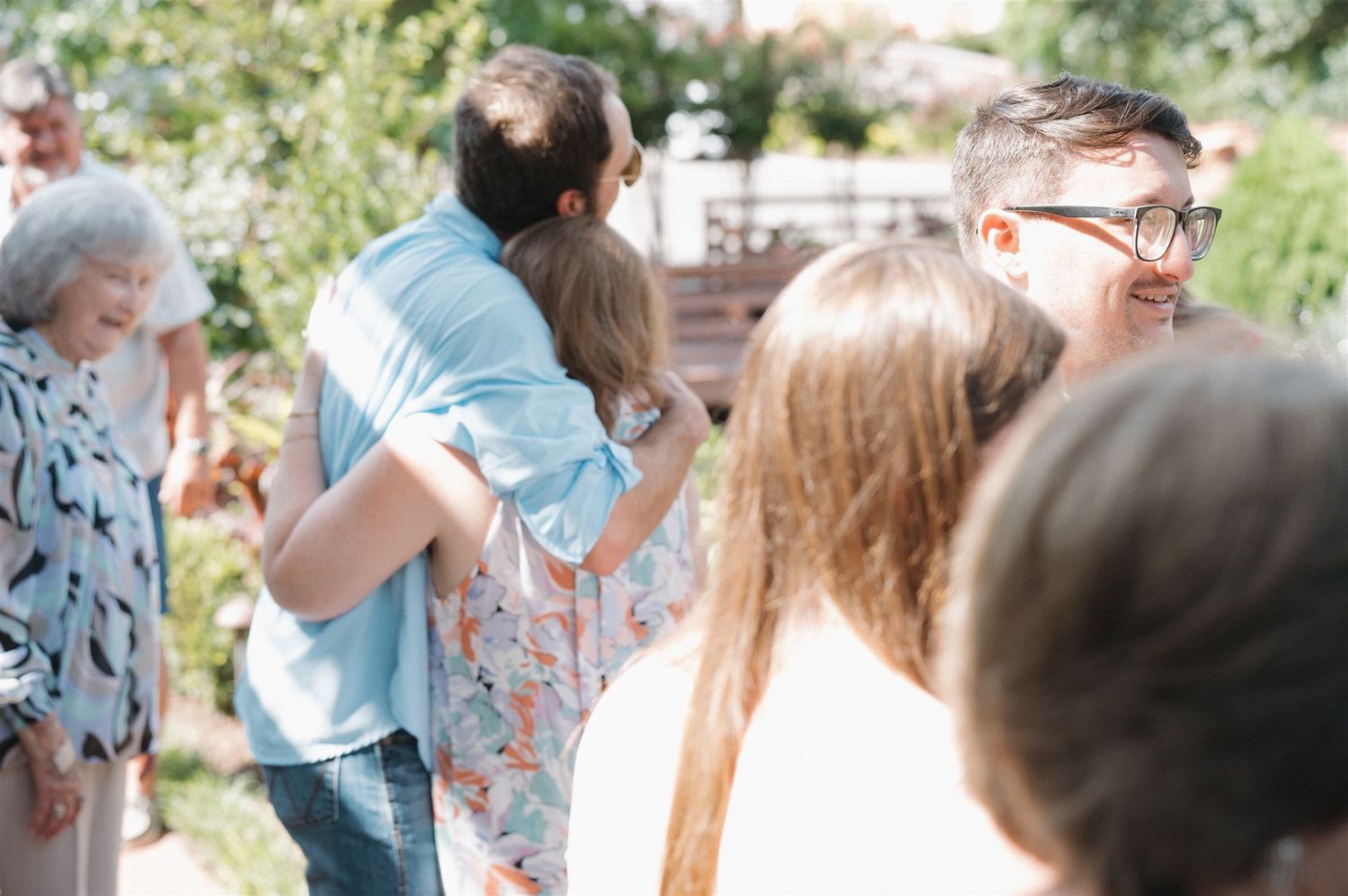 A group of people gathered outdoors in a sunny garden setting having conversations in casual summer attire.