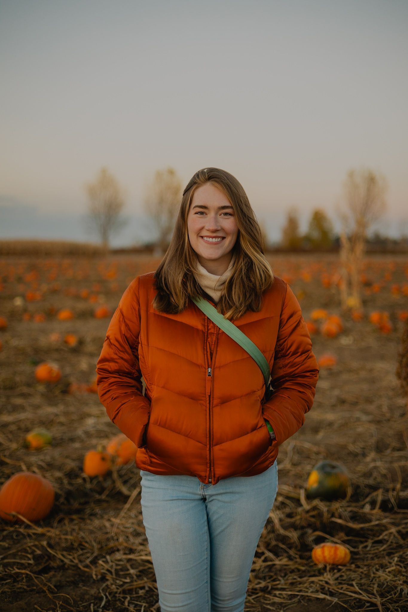 Woman in bright orange puffy jacket standing in pumpkin patch during autumn sunset.