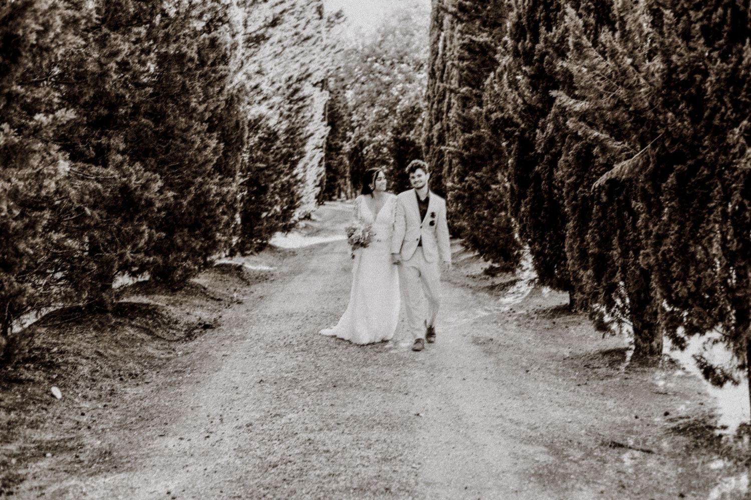 A black and white photo of a couple walking along a cypress-lined path in wedding attire.