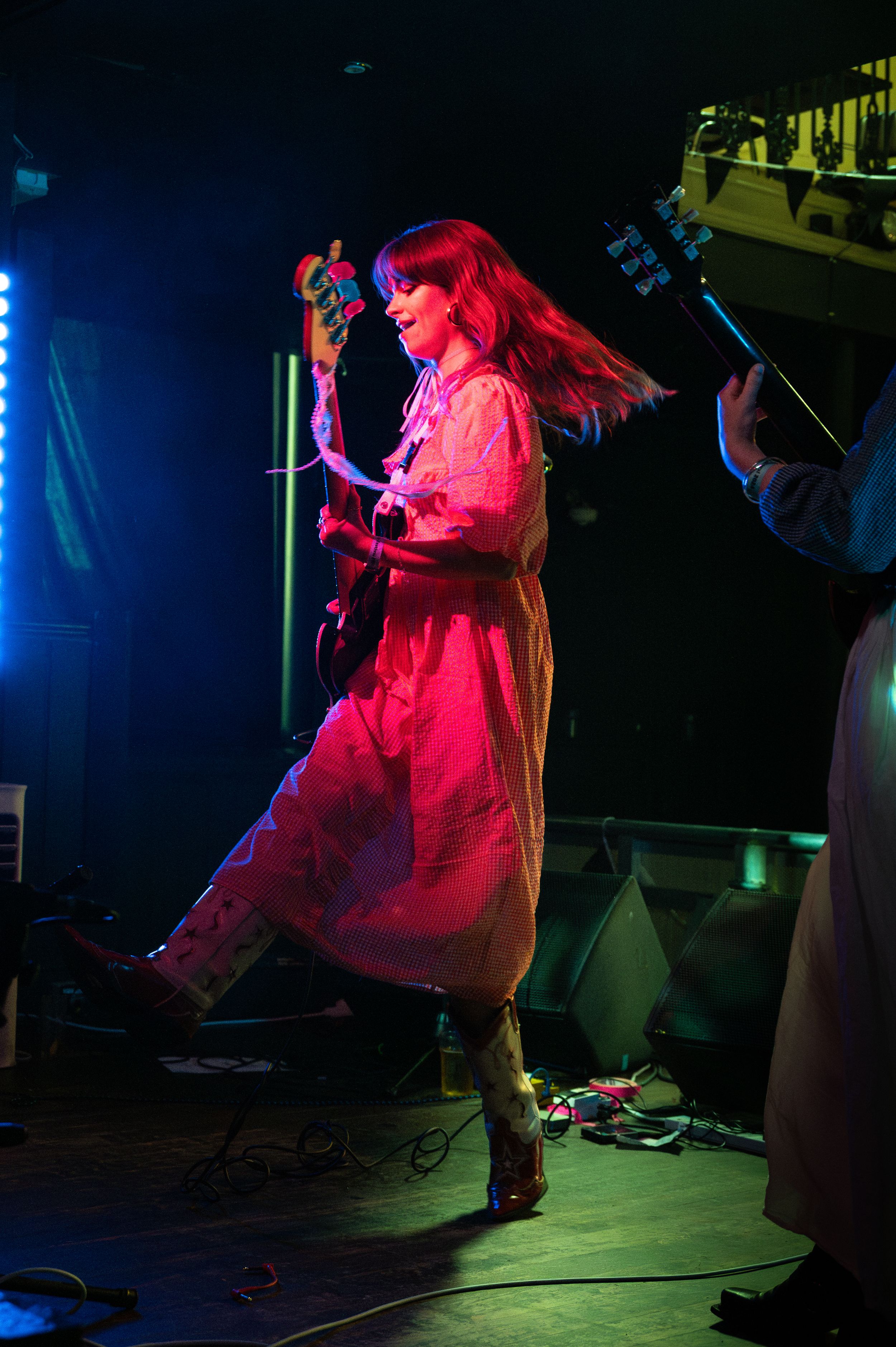 Performer in flowing dress plays guitar under dramatic red stage lighting.