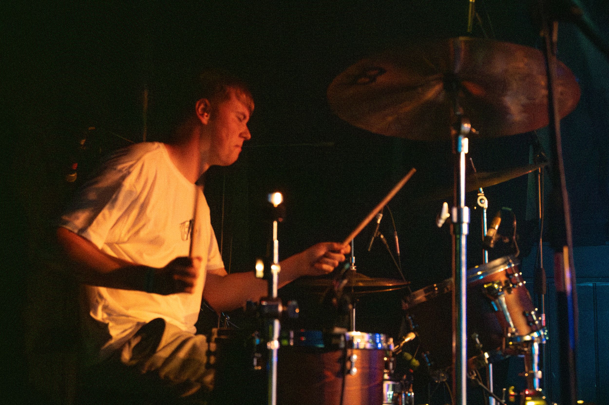 Drummer plays kit under warm stage lighting during live performance.