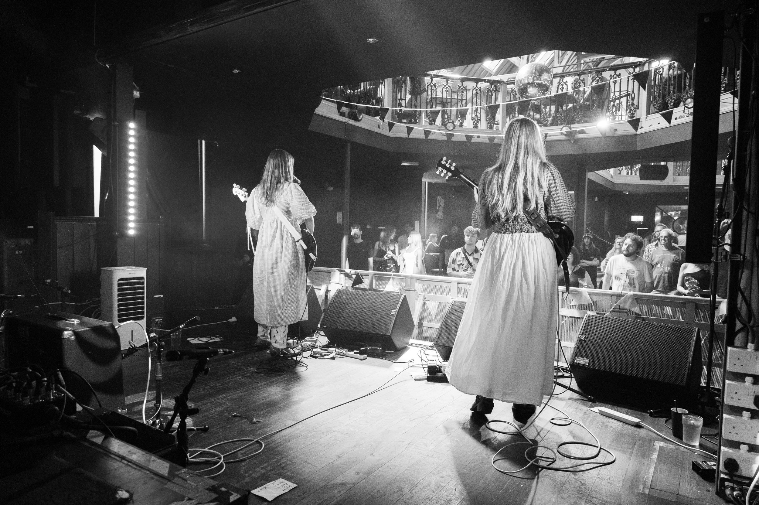 Black and white photo of performers in white dresses on a dark stage with drum kit and equipment visible.