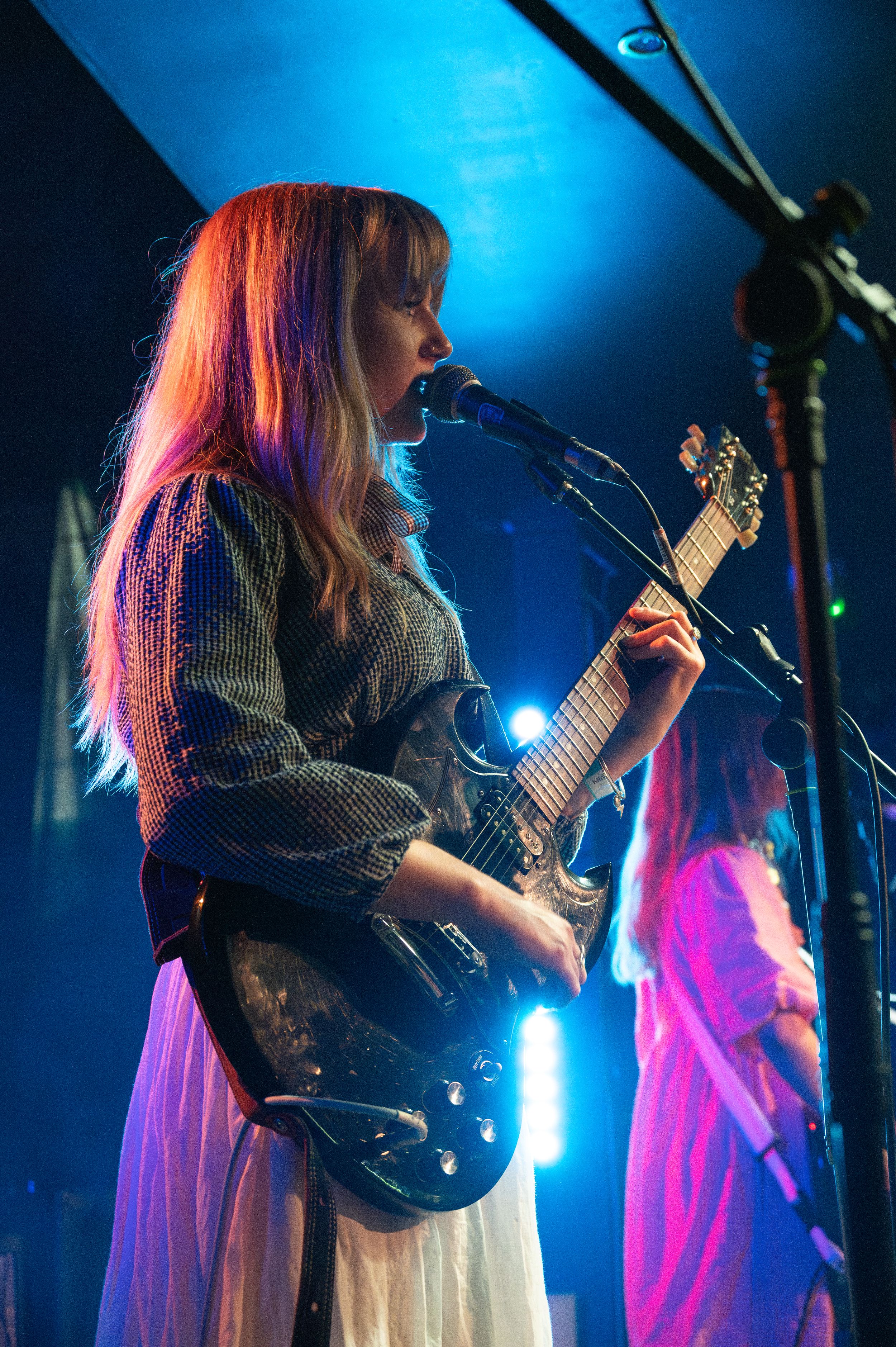 Musician in white dress plays electric guitar against blue-lit stage backdrop.