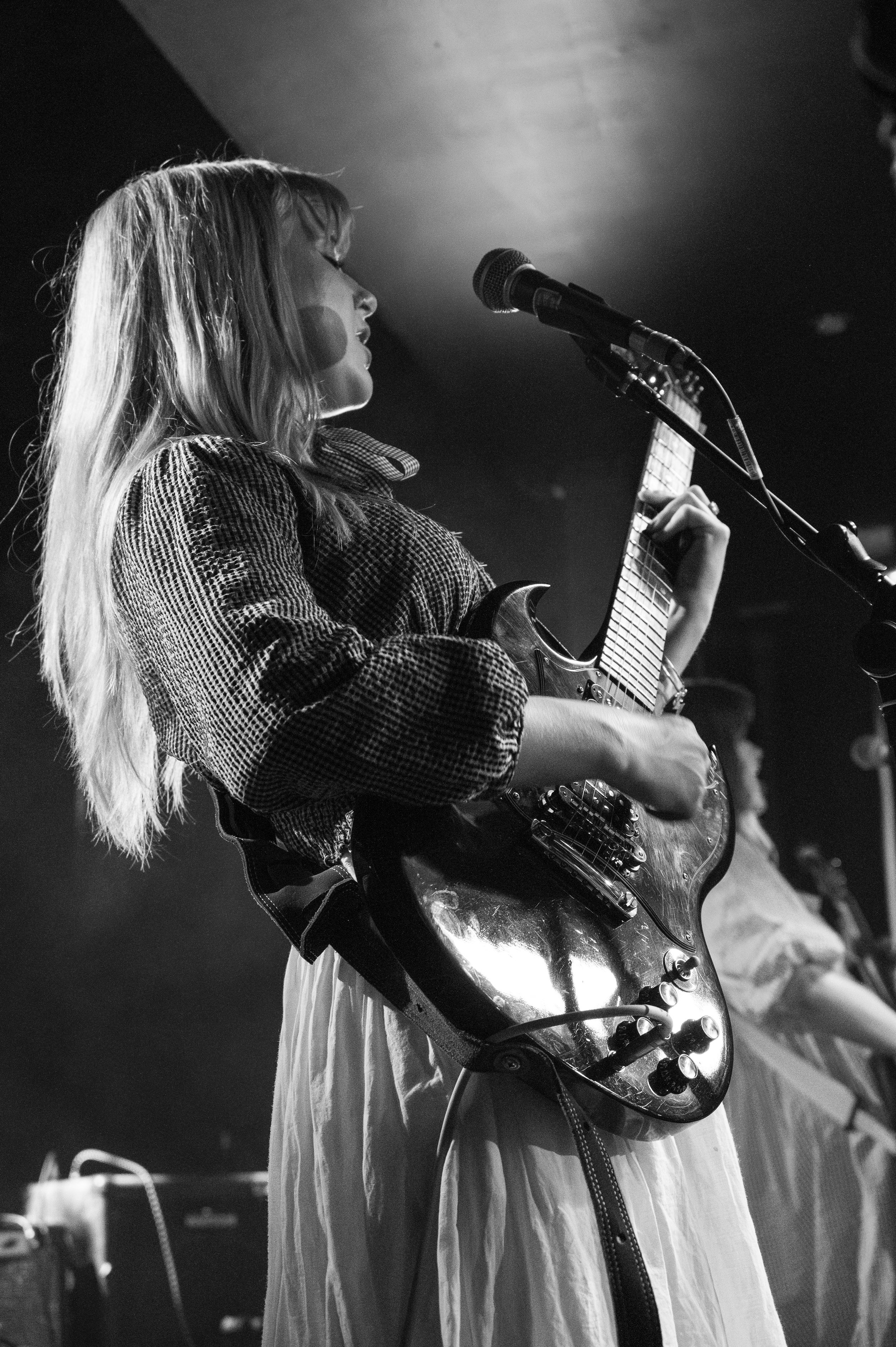 Black and white concert photo of guitarist in sweater and white dress performing on stage.