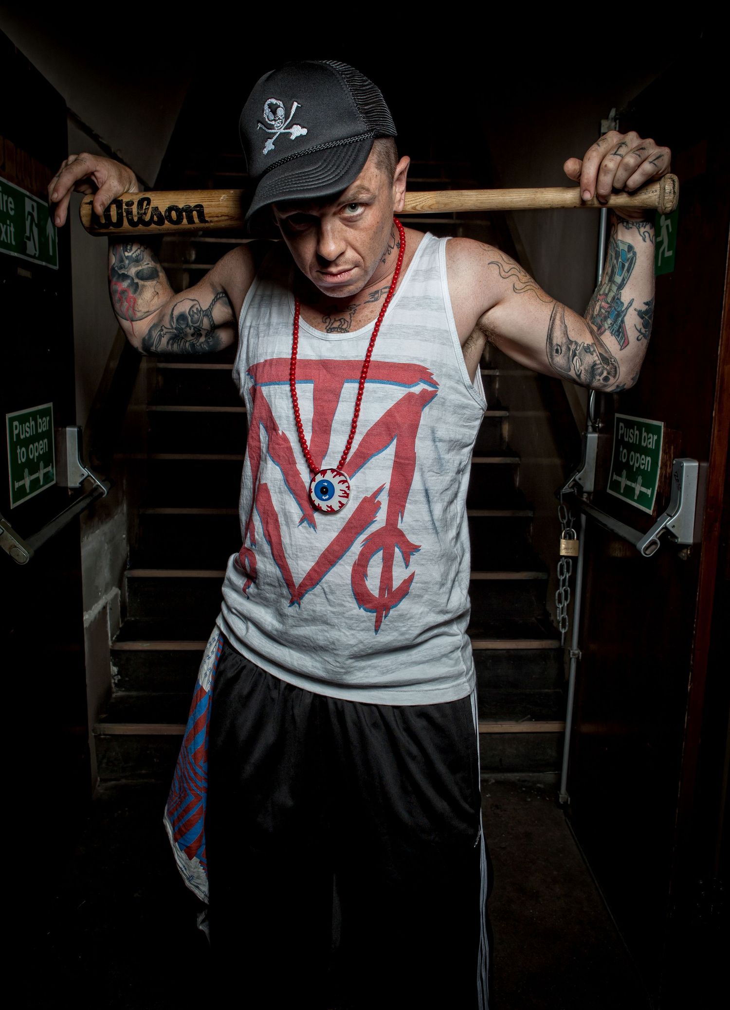 Tattooed man in gray tank top and cap poses with baseball bat on shoulders near dark staircase.