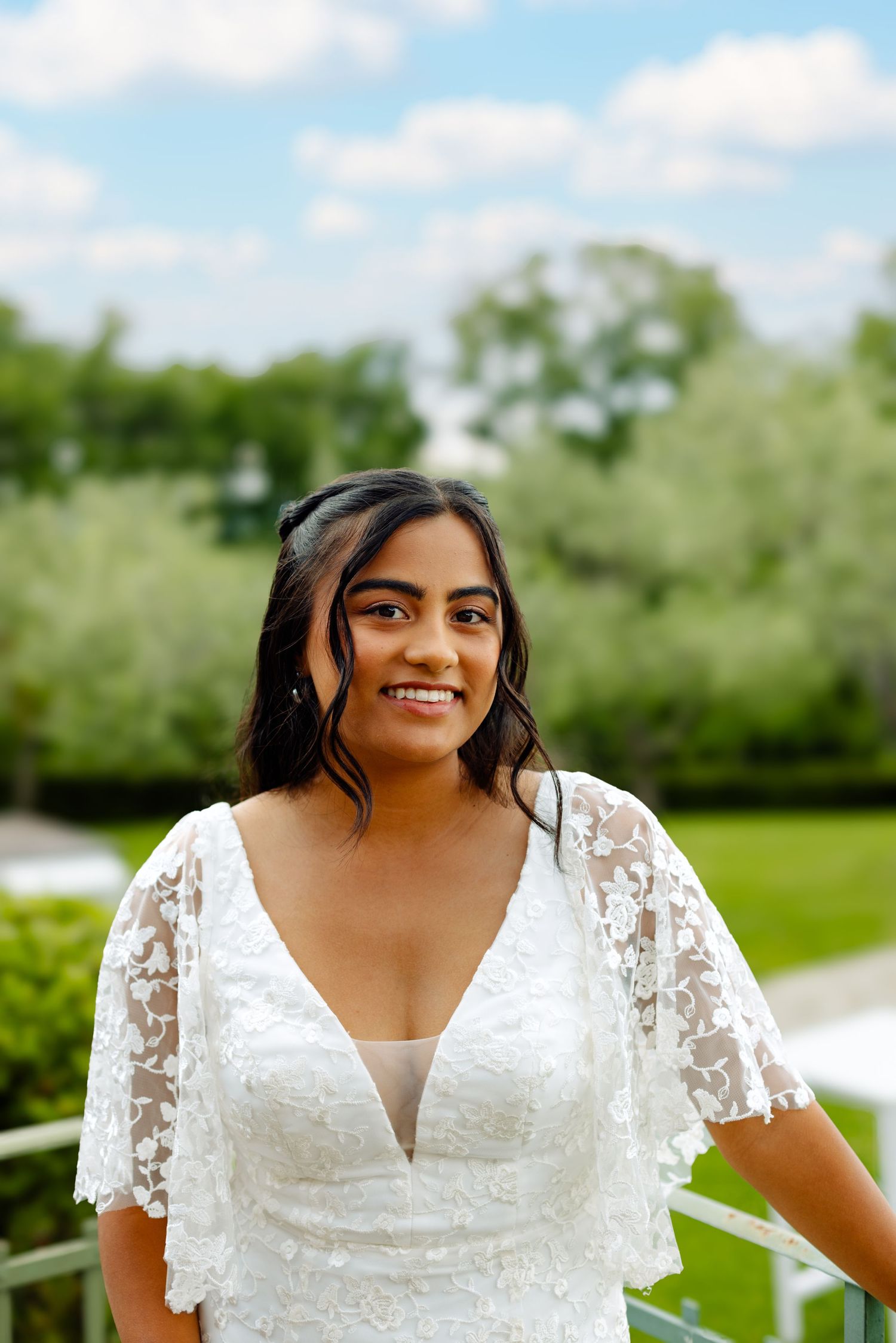 A bride in a beautiful lace dress smiles radiantly outdoors on her wedding day.