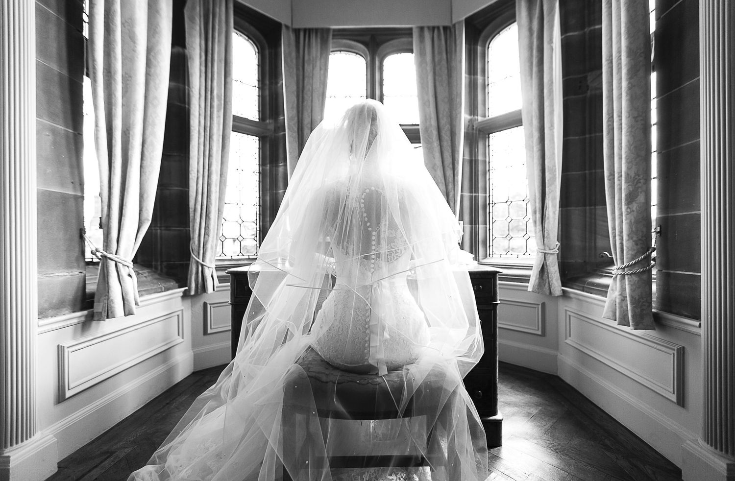 Bride in white wedding gown and veil sitting by large windows in elegant black and white photo.