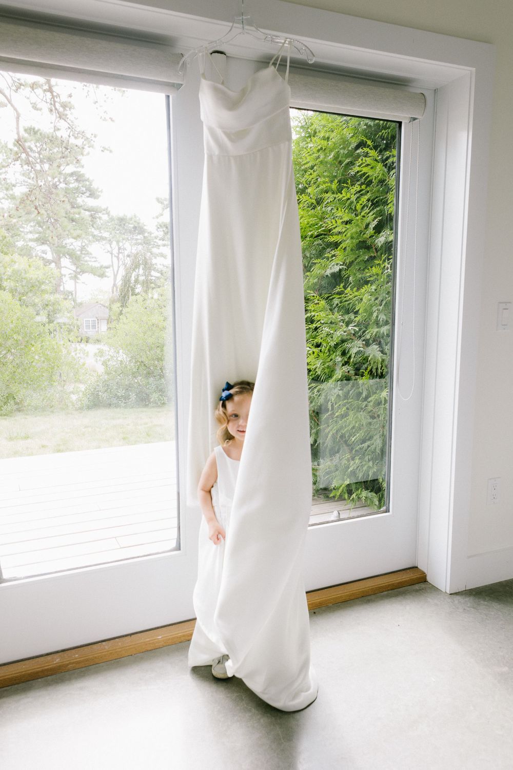 White wedding dress hanging by a bright window with sunlight streaming in.