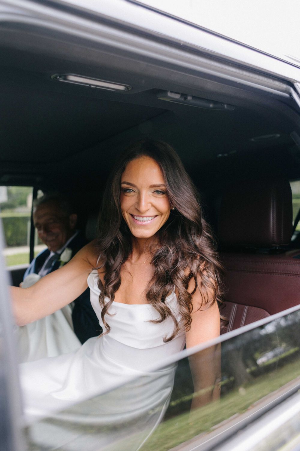 A bride in a white gown smiles radiantly from inside a luxury car.