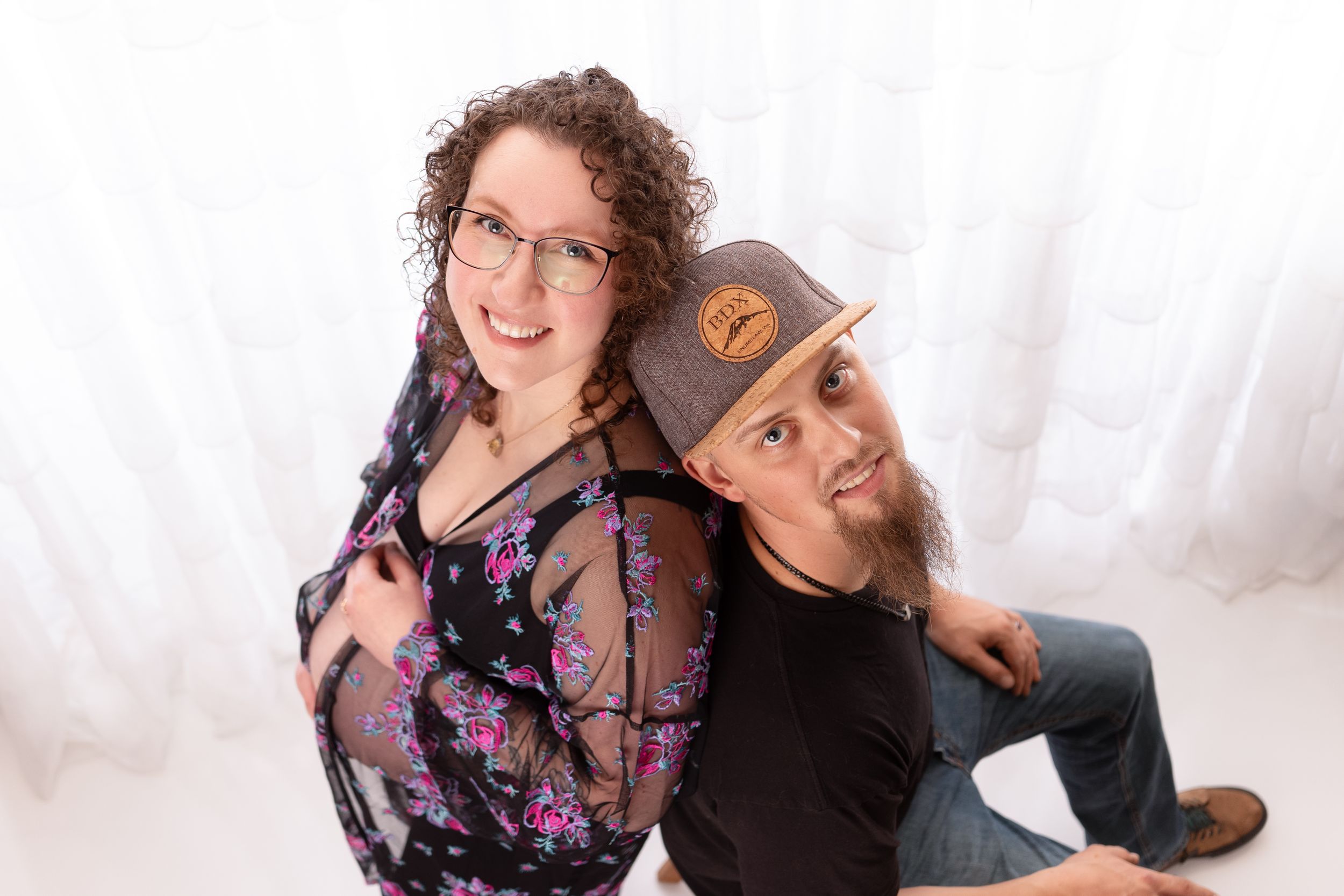 Couple poses playfully for maternity photo against white backdrop.