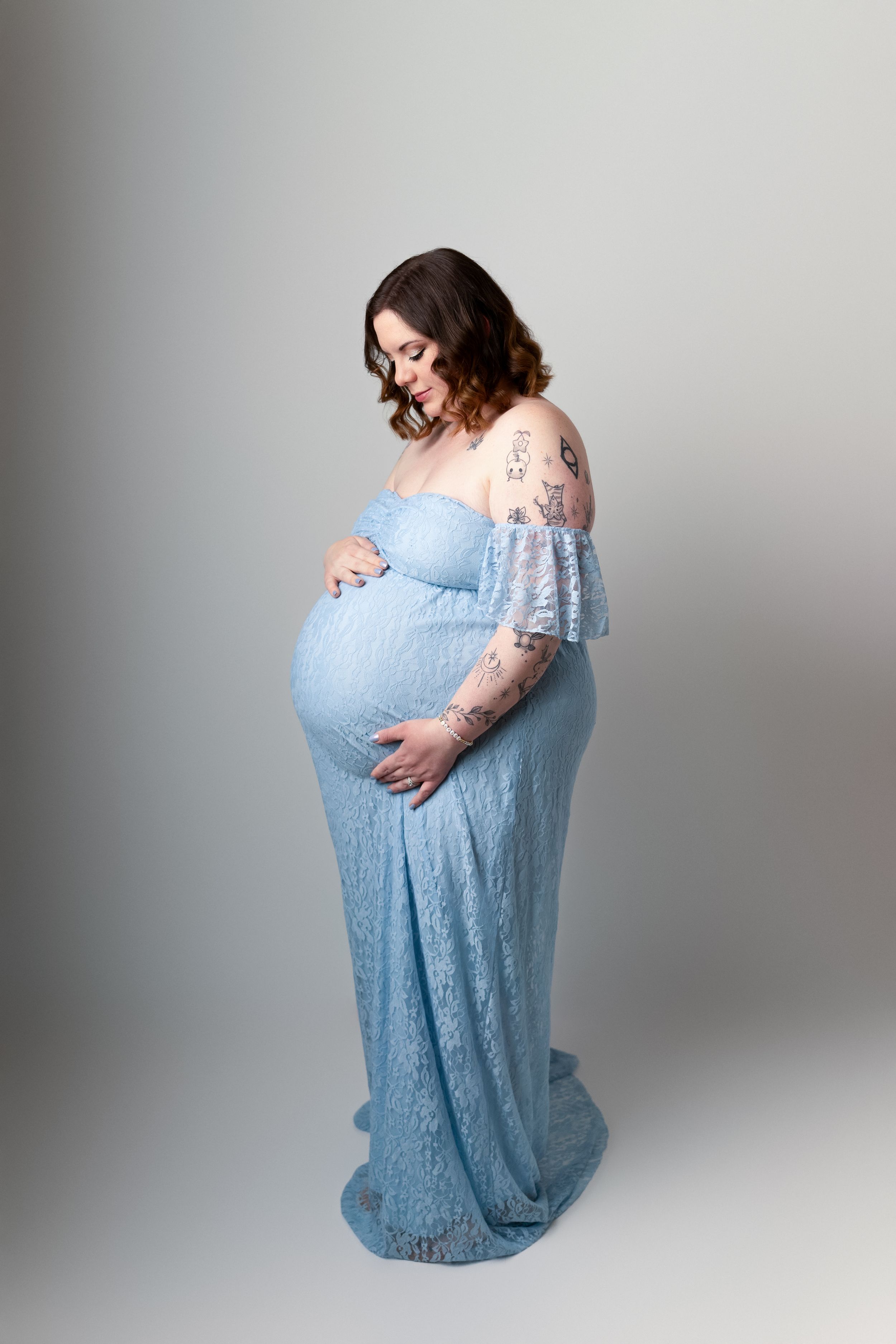 Woman in flowing blue lace dress cradles her pregnant belly against grey backdrop. Maternity photography session in Enumclaw, Washington by