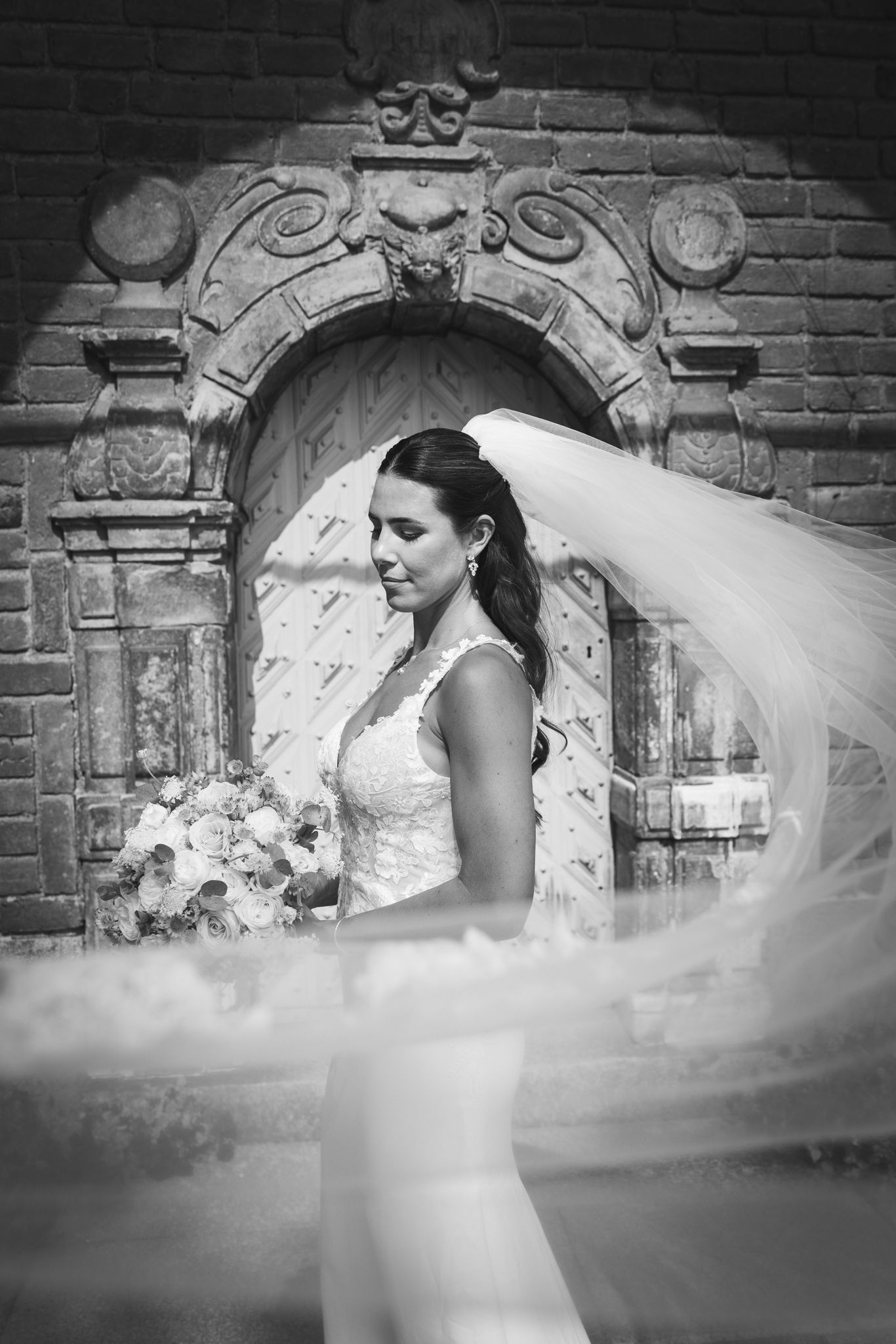 A flowing white wedding veil creates an ethereal effect against a rustic brick wall backdrop in black and white.