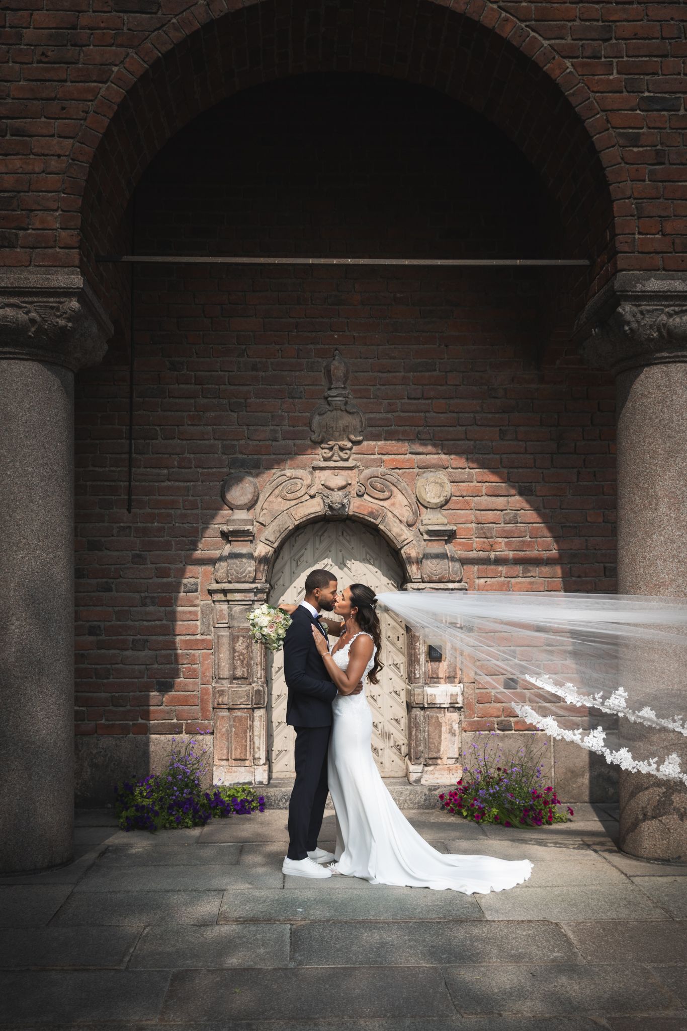 Romantic wedding photos captured under brick archways with a dramatic flowing veil catching the light.