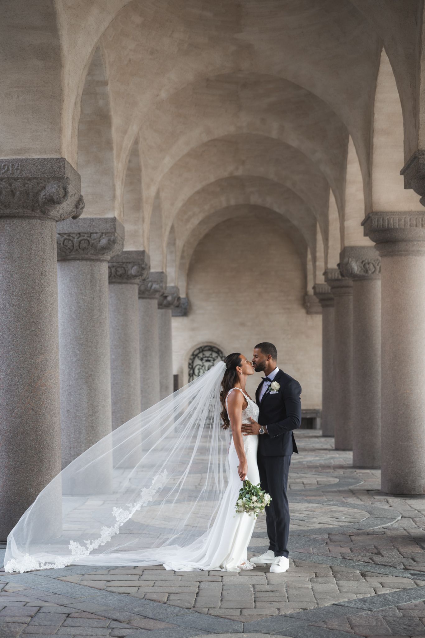Elegant wedding couple photographed in a stone colonnade with a flowing cathedral veil creating a romantic atmosphere.