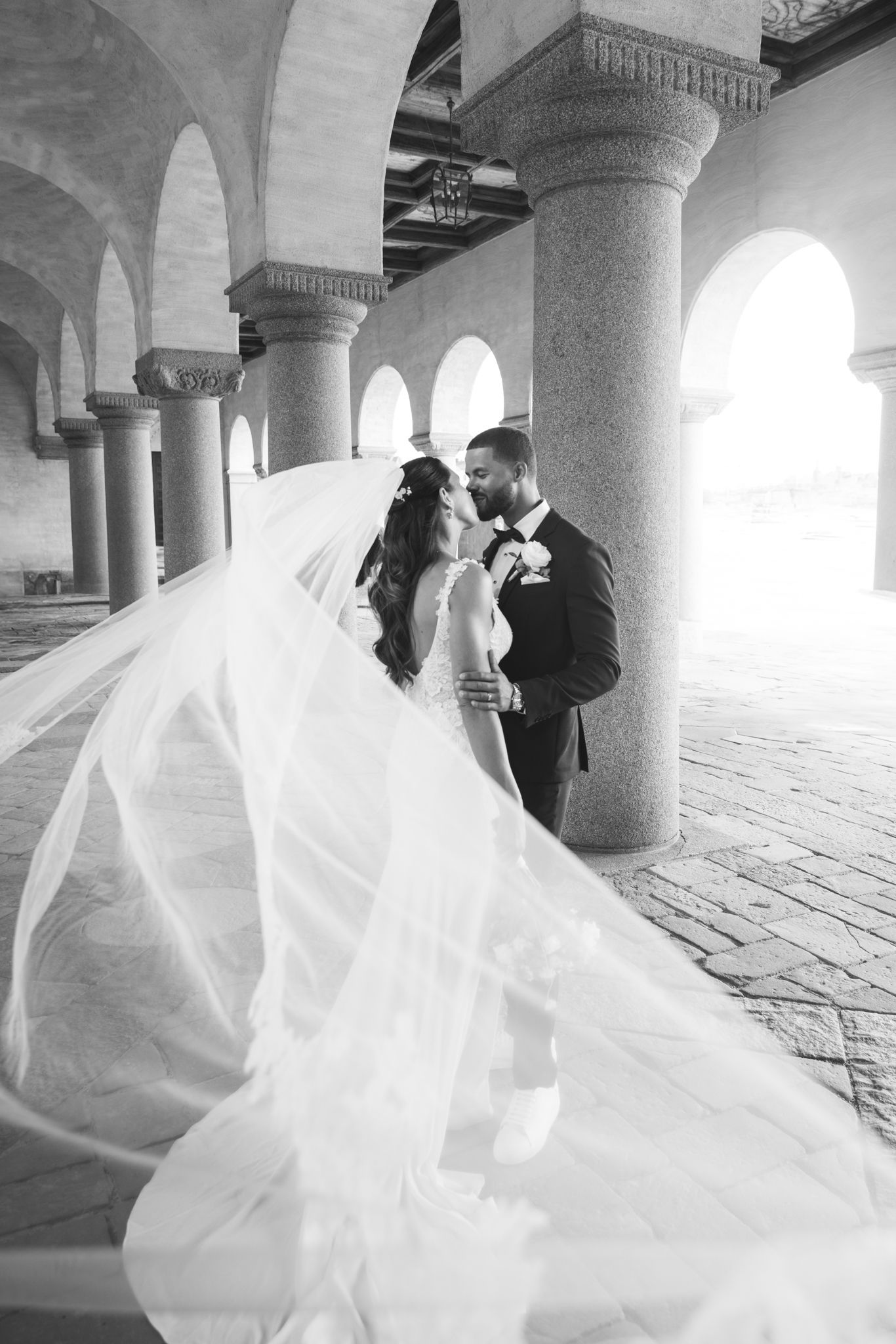 A romantic black and white wedding photoshoot in a columned courtyard with a flowing veil catching the light.