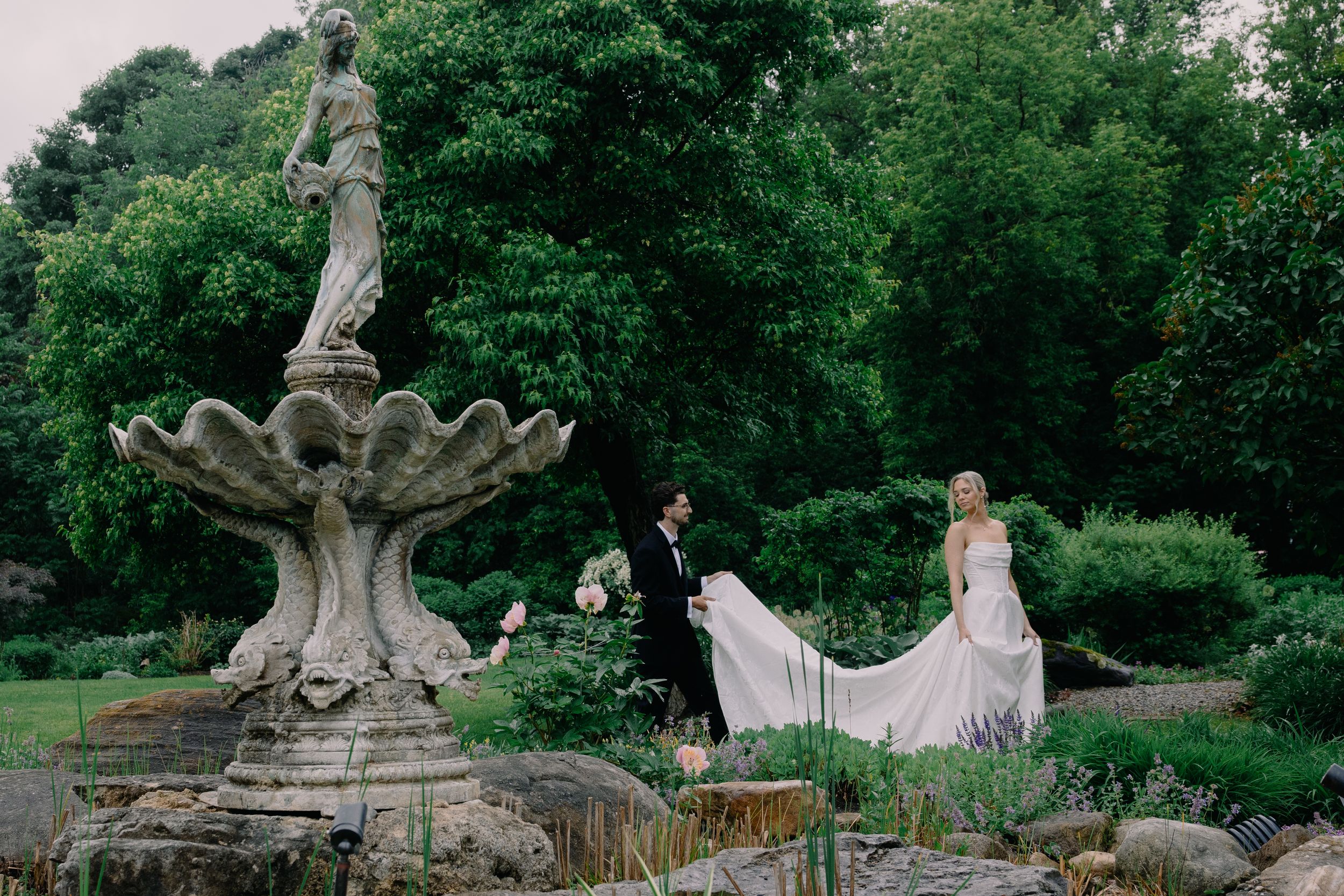 Bride and groom walk past an ornate stone fountain in a lush garden wedding setting.