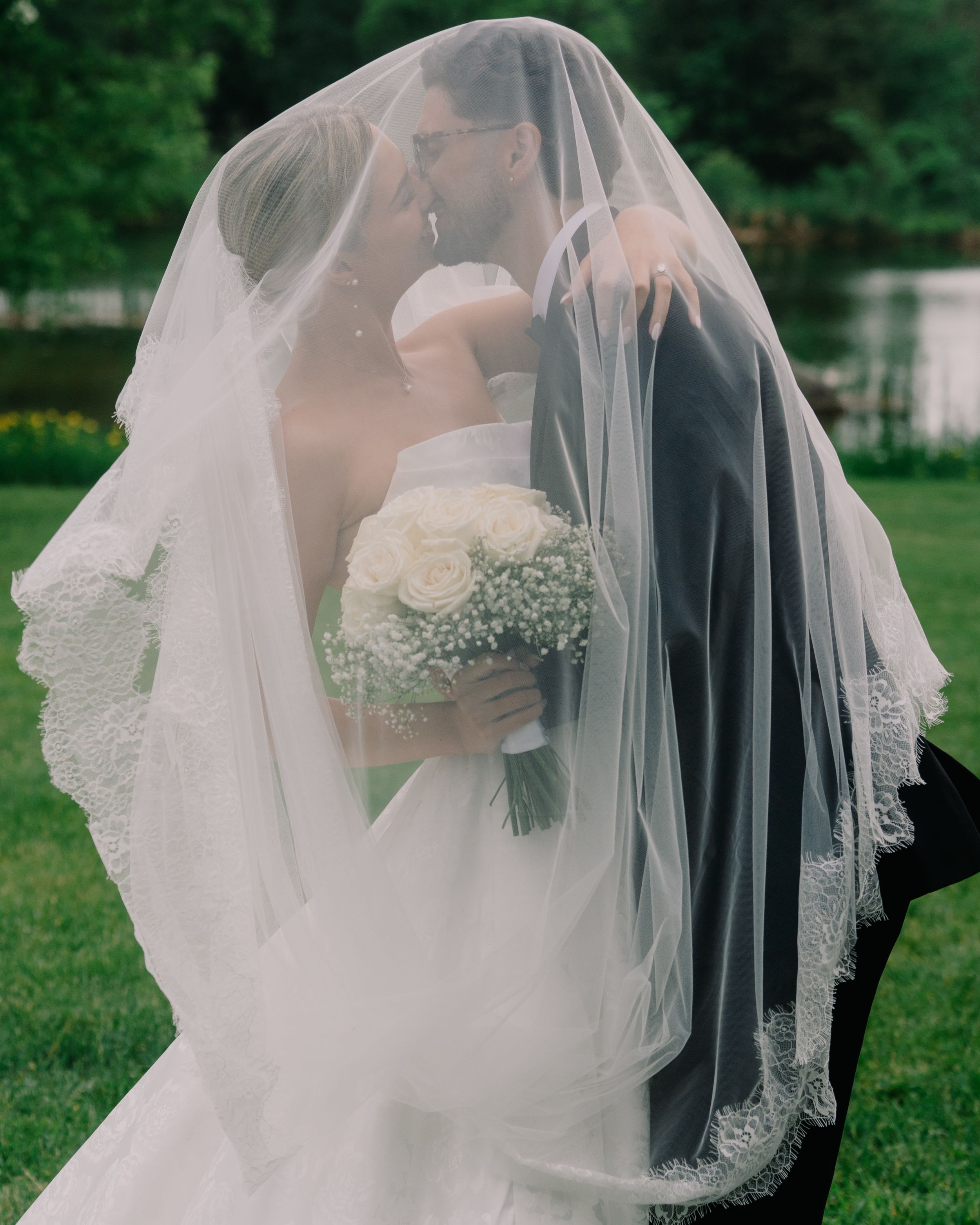 Bride and groom sharing a romantic kiss under a white lace wedding veil outdoors, bride holding a white rose bouquet.