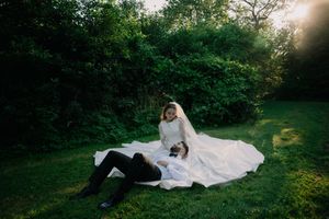 Bride in flowing white gown sits with groom on lush green grass during golden hour outdoor wedding portrait.