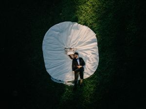 Aerial view of a wedding couple lying on a white bridal gown spread on lush green grass.
