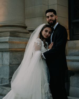 A bride in a white gown and veil embraces a groom in a black tuxedo on stone steps in a romantic wedding portrait.