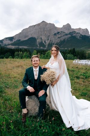 A couple in wedding attire sits together on rocks with majestic mountain peaks rising in the background.