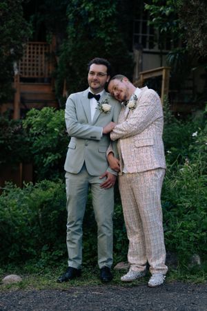 A tender wedding moment captured in a garden setting with one person resting their head on another's shoulder.