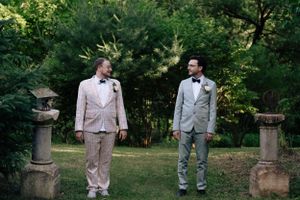 Two people in light-colored suits share a moment in a garden setting between stone pillars.