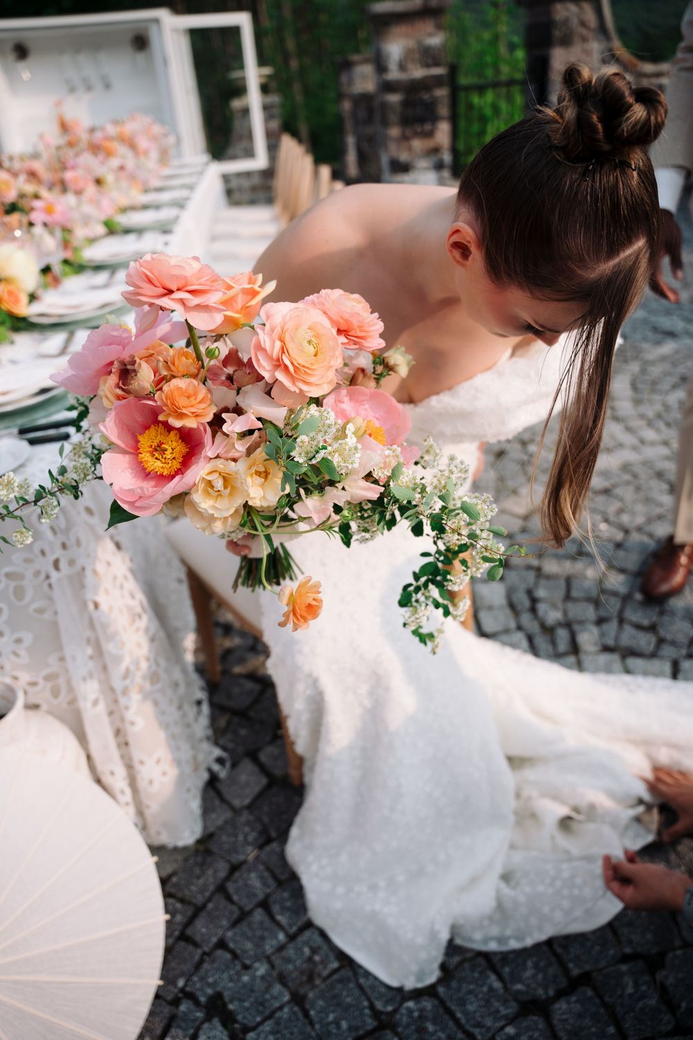 A bride in a white dress holds a peach and coral floral bouquet while walking on cobblestone streets.