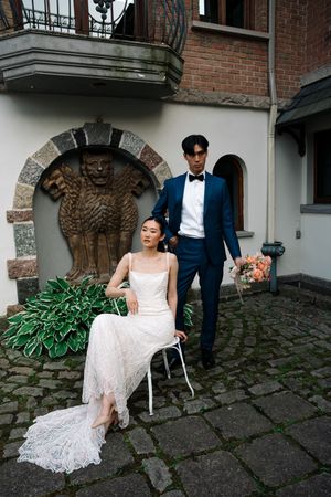 A wedding couple poses in front of a stone archway with ornate architectural details and a lush garden setting.
