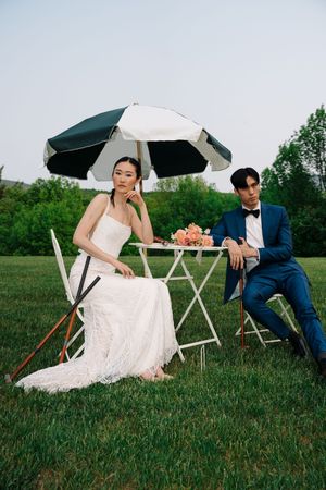 A couple in wedding attire poses playfully with black umbrellas while sitting on grass during an outdoor photoshoot.