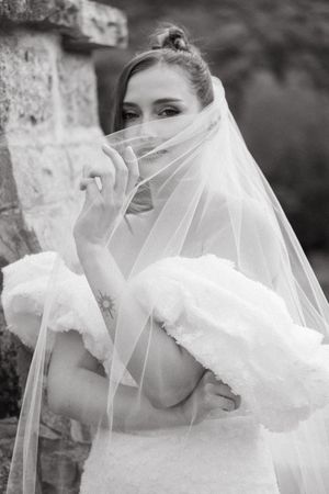 Black and white portrait of a bride in a flowing veil and wedding dress posing against a stone wall.