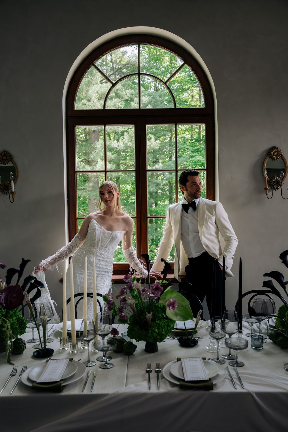 Elegant wedding reception table settings with white linens and candles by arched windows overlooking a garden.