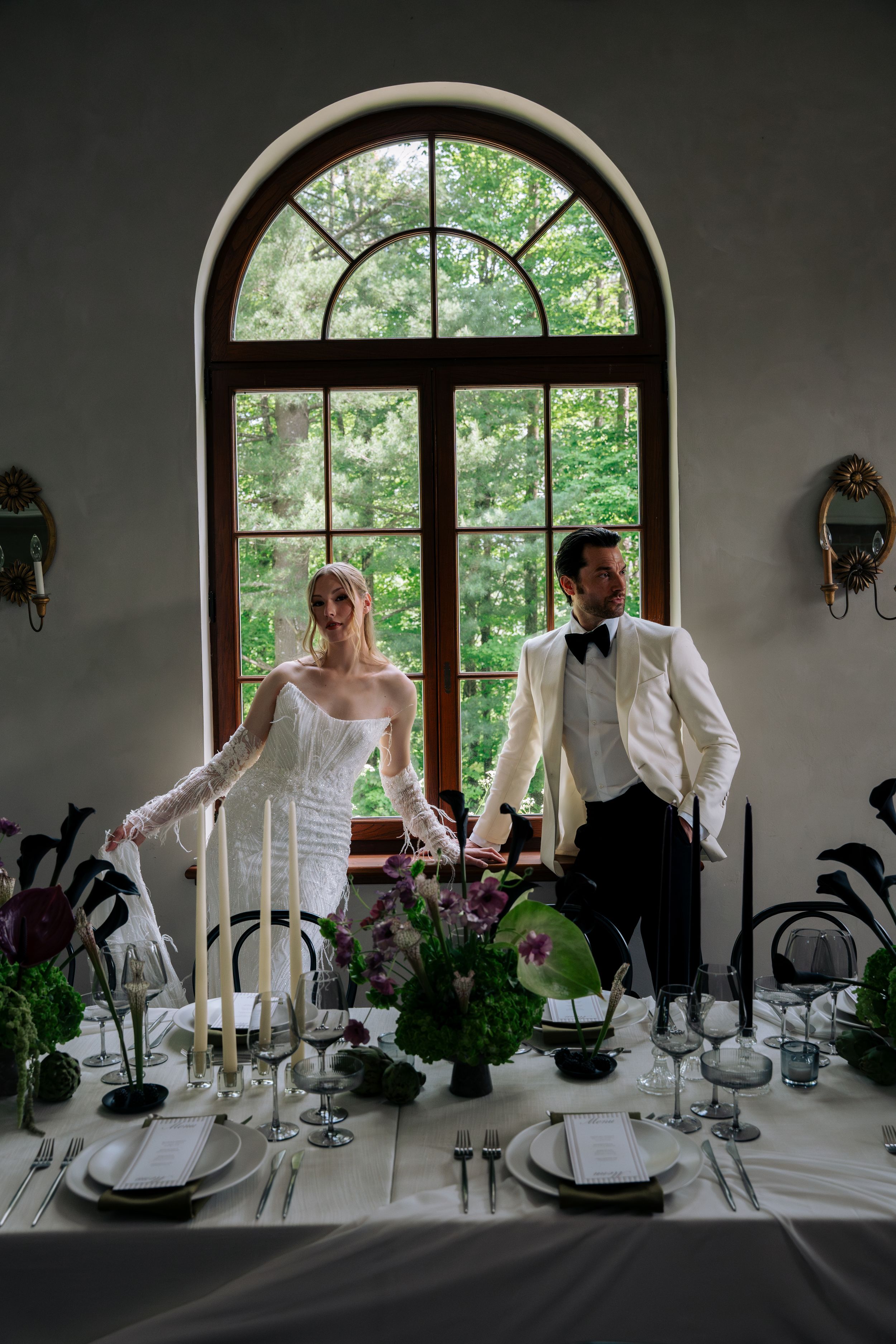 Elegant wedding reception table settings with white linens and candles by arched windows overlooking a garden.