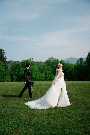 Newlyweds walk across a lush green field with mountains and trees in the background on their wedding day.