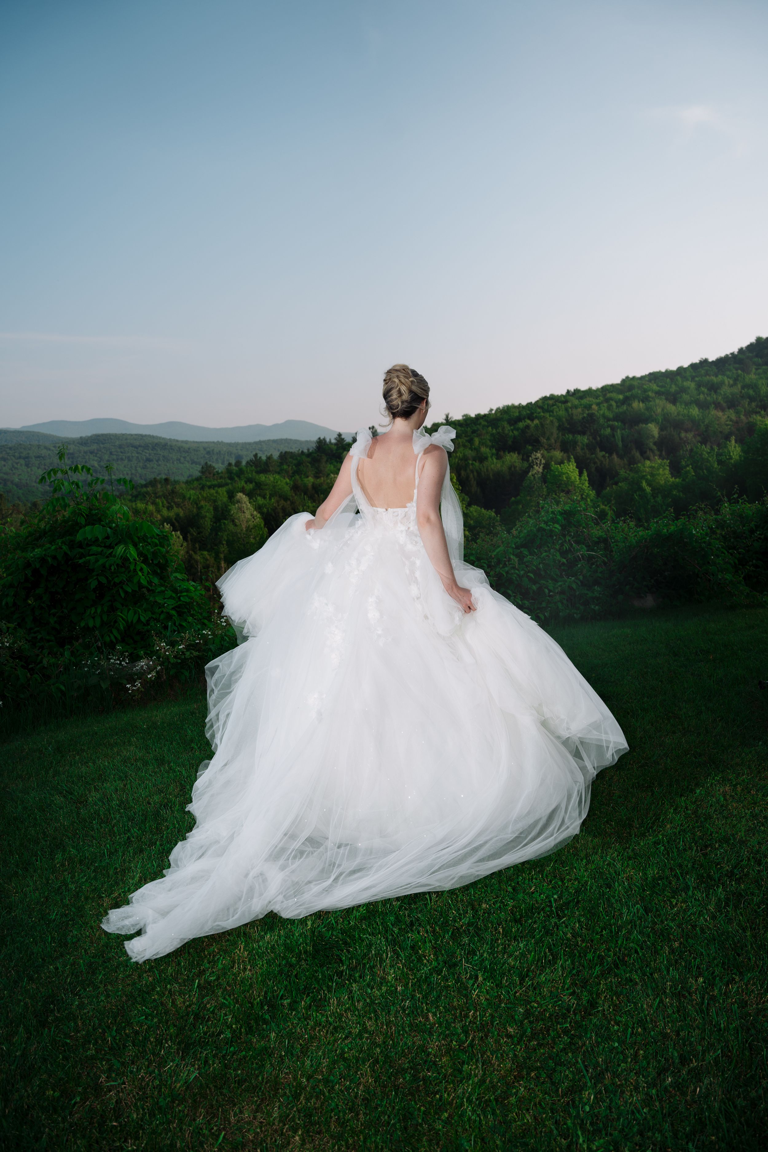 A flowing white wedding dress billows gracefully across a green hillside with mountains in the background at dusk.