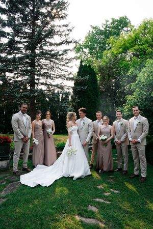 Wedding party in tan suits and mauve dresses poses on a grassy lawn with evergreen trees in the background.