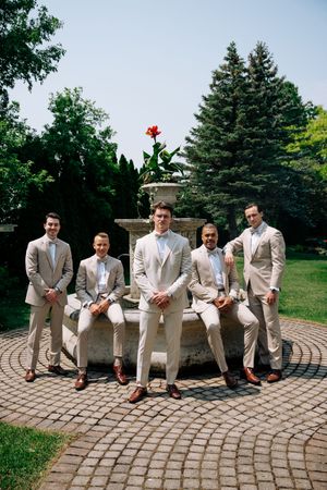 Groomsmen in light beige suits pose around an ornate fountain in a garden setting with evergreen trees.