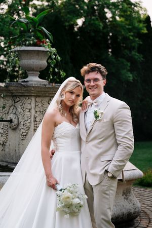 Romantic wedding portraits of a couple in white and beige attire posing by ornate stone pillars in a garden.
