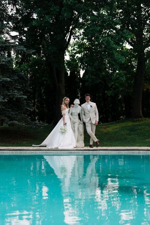 Wedding couple poses for photos beside a bright turquoise swimming pool surrounded by lush trees.