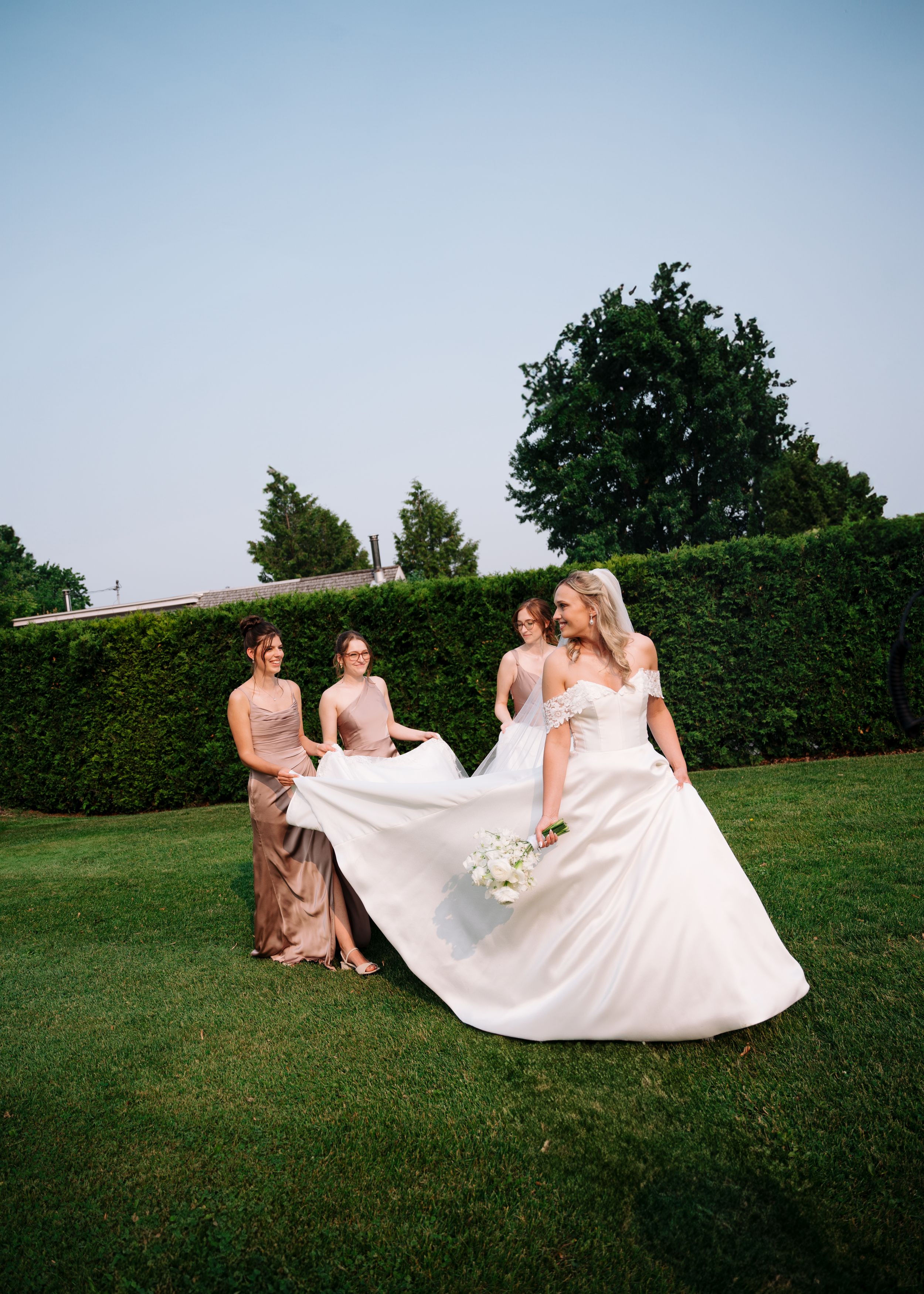 Wedding party members in blush pink dresses help arrange a flowing white wedding gown on a manicured lawn.