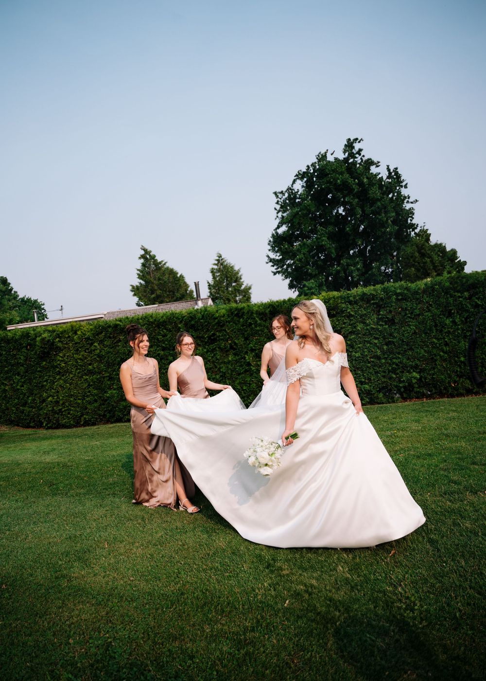 Wedding party members in blush pink dresses help arrange a flowing white wedding gown on a manicured lawn.