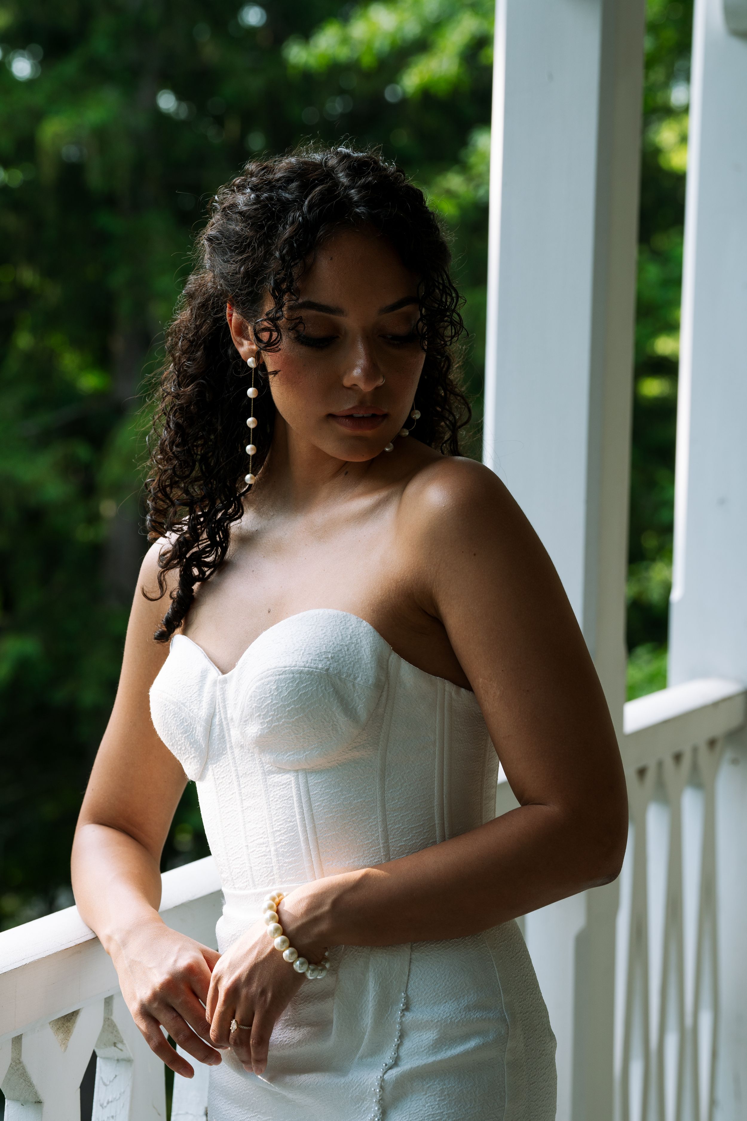 Woman in white strapless dress stands thoughtfully on a white porch with greenery in background.