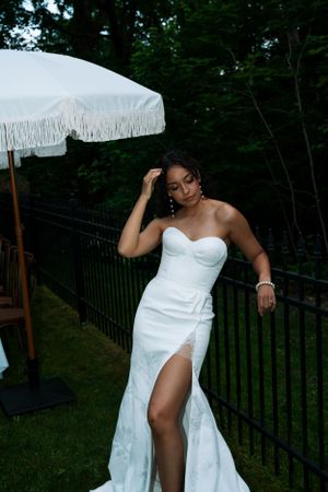 Elegant bride in fitted white gown with thigh slit poses under white umbrella outdoors.
